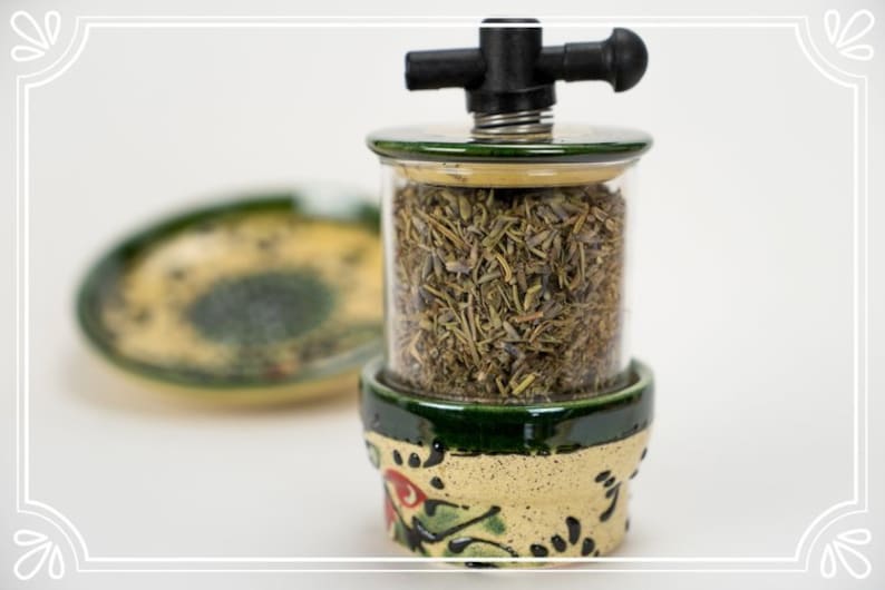 May include: A ceramic herb grinder with a green and yellow floral design. The grinder has a clear glass jar filled with dried herbs and a black plastic handle on top.