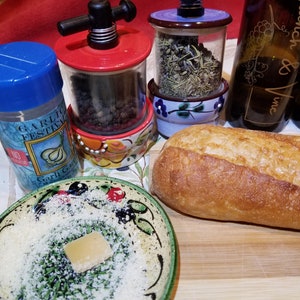 May include: A close-up of a wooden cutting board with a loaf of bread, a small bowl of grated cheese, and three ceramic spice grinders. The grinders are filled with different spices, including black peppercorns, herbs, and garlic. The grinders are decorated with floral patterns and have a rustic, farmhouse style.