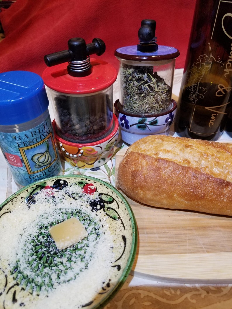 May include: A close-up of a wooden cutting board with a loaf of bread, a small bowl of grated cheese, and three ceramic spice grinders. The grinders are filled with different spices, including black peppercorns, herbs, and garlic. The grinders are decorated with floral patterns and have a rustic, farmhouse style.