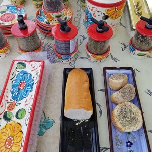 May include: A collection of ceramic spice jars with red lids and floral designs. The jars are filled with various spices, including black peppercorns, herbs, and dried oregano. The jars are displayed on a white tablecloth with a floral pattern. There are also two ceramic serving dishes, one with a black background and the other with a blue background. The dishes are filled with bread and garlic bread.