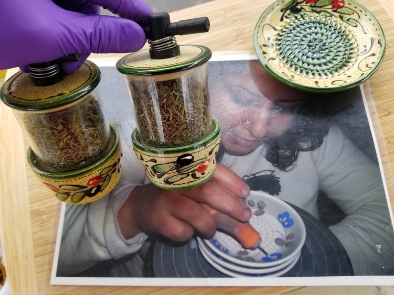 May include: Two ceramic spice grinders with clear glass jars filled with dried herbs. The grinders have a green and yellow floral design with black accents. The grinders are on a wooden surface.