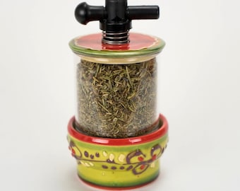 May include: A clear glass herb grinder with a black handle and a red and green decorative base. The grinder is filled with dried herbs. The base has a yellow background with red and green accents.