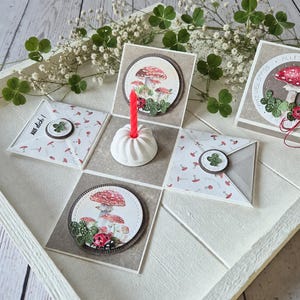 Könnte beinhalten: Eine quadratische Geschenkbox aus Papier, deren Deckel sich aufklappt und einen weißen, geriffelten Kerzenhalter mit einer roten Kerze enthüllt. Die Paneele zeigen Illustrationen von roten Pilzen, grünem Klee und Marienkäfern sowie den Text AUF DICH.