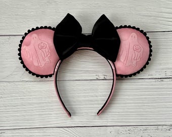 May include: Pink Halloween-themed headband with black trim and a large black bow. The round ears feature a ghost and pumpkin design. The headband is a festive accessory for Halloween.