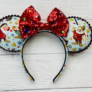 May include: Red sequin bow headband with a blue and white fabric design featuring the seven dwarfs from Disney's Snow White and the Seven Dwarfs.