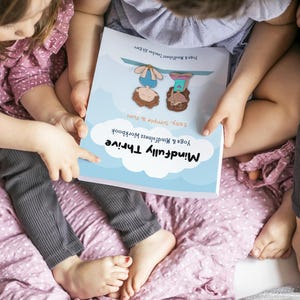 K&ouml;nnte beinhalten: Zwei Kinder sitzen auf einer rosa Decke und betrachten ein Buch mit dem Titel "Mindfully Thrive: Yoga & Mindfulness Workbook, Easy, Simple & Fun".