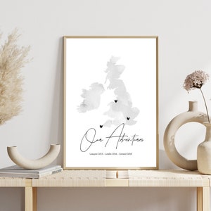 May include: A black and white watercolour print of the United Kingdom with three black heart markers. The text "Our Adventures" is written in cursive below the map with the dates "Liverpool 2013 - London 2014 - Cornwall 2018".