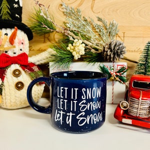 Pode incluir: Uma caneca de cerâmica azul com esmalte branco salpicado. A caneca tem o texto "Let it snow" escrito três vezes em letras brancas.
