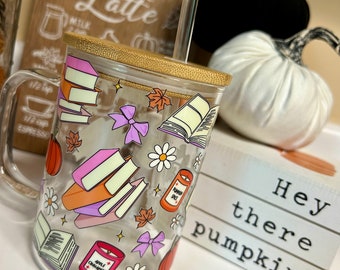 May include: A clear glass mug with a bamboo lid and a straw. The mug has a fall-themed design with books, candles, pumpkins, and bows. The text "Hey there pumpkin" is visible on a white sign in the background.