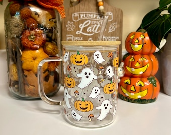 May include: A clear glass mug with a bamboo lid and a straw. The mug has a Halloween theme with a pattern of orange pumpkins and white ghosts. The mug is sitting on a white surface with a jar of fall decorations and a wooden sign that says "Pumpkin Spice Latte" in the background.