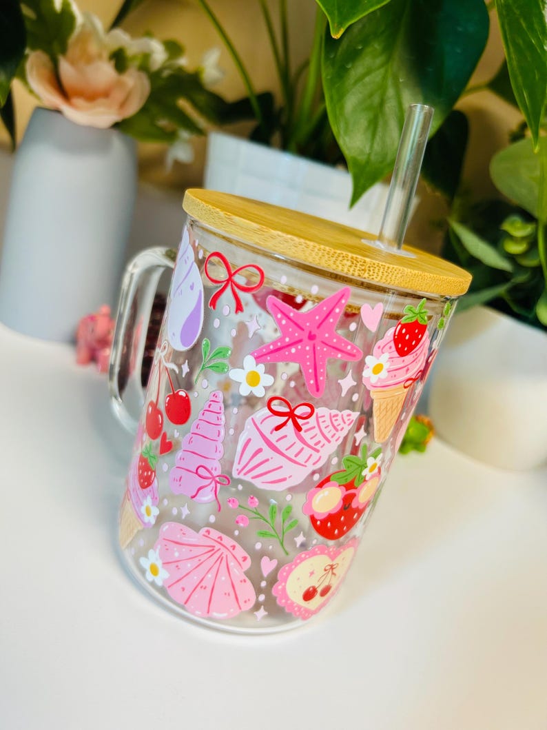 Peut inclure: Mug en verre transparent avec couvercle et paille en bambou, d&eacute;cor&eacute; d'illustrations roses et rouges sur le th&egrave;me de l'&eacute;t&eacute;. Le motif comprend une &eacute;toile de mer, des coquillages, des cornets de glace, des cerises et des fraises.