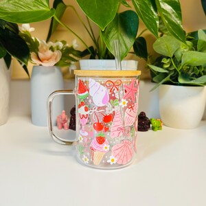 Peut inclure: Un mug en verre transparent avec une anse, d&eacute;cor&eacute; d'illustrations sur le th&egrave;me de l'&eacute;t&eacute;. Le motif comprend des images de past&egrave;que, de cr&egrave;me glac&eacute;e, de cerises, de coquillages et d'&eacute;toiles de mer. Le mug a un couvercle en bambou et une paille transparente.