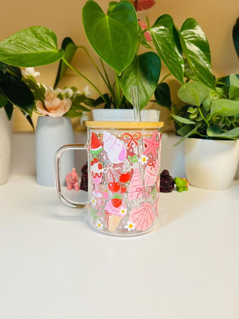 Peut inclure: Un mug en verre transparent avec une anse, d&eacute;cor&eacute; d'illustrations sur le th&egrave;me de l'&eacute;t&eacute;. Le motif comprend des images de past&egrave;que, de cr&egrave;me glac&eacute;e, de cerises, de coquillages et d'&eacute;toiles de mer. Le mug a un couvercle en bambou et une paille transparente.