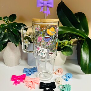 Peut inclure: Un mug en verre transparent avec un couvercle en bambou et un motif floral, avec un dessus de paille en forme de nœud papillon violet. Plusieurs accessoires en forme de nœud papillon colorés en rose, bleu et noir sont affichés. Le mug est entouré de plantes en pot.