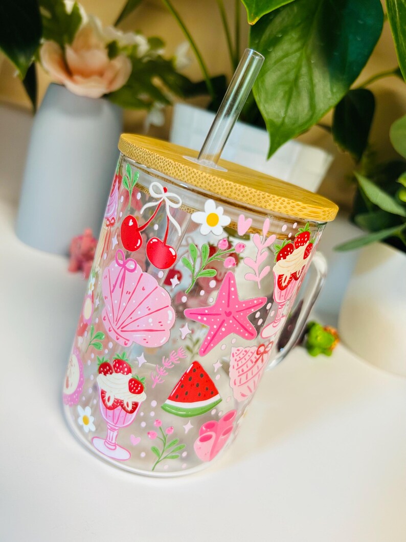 Peut inclure: Un gobelet en verre transparent avec un couvercle en bambou et une paille en verre. Le gobelet est d&eacute;cor&eacute; d'illustrations roses et rouges sur le th&egrave;me de l'&eacute;t&eacute;, notamment des cerises, un coquillage, une &eacute;toile de mer et une tranche de past&egrave;que.