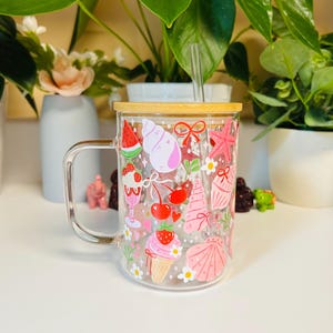 Peut inclure: Mug en verre transparent avec un couvercle en bambou et une paille transparente. Le mug est d&eacute;cor&eacute; d'illustrations color&eacute;es sur le th&egrave;me de l'&eacute;t&eacute;, notamment des tranches de past&egrave;que, des coquillages, des cerises et des cornets de glace. Le mug a une anse.
