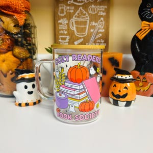 May include: A clear glass mug with a gold lid and straw, featuring the text "COZY READERS BOOK SOCIETY." The mug is decorated with pumpkins, books, ghosts, and leaves. The background includes autumn-themed decorations.