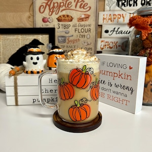 May include: A glass mug with a pumpkin spice latte and whipped cream. The mug has a design of orange pumpkins with green stems. The mug is sitting on a wooden coaster. The background includes a sign that says "If loving pumpkin spice is wrong, I don't wanna be right."