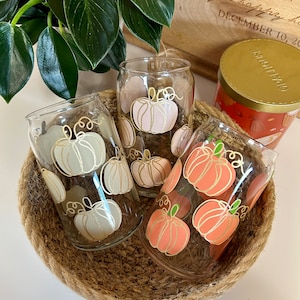 May include: Three glass tumblers with pumpkin designs. The tumblers are in a woven basket. The pumpkins are painted in shades of pink, peach, and white.