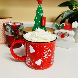 May include: Red ceramic mug with white Christmas tree designs and a white whipped cream topping. The mug is sitting on a white surface with a wooden base and a green Christmas tree decoration behind it.