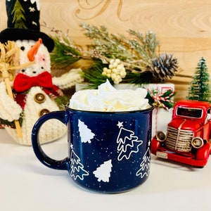 May include: A blue ceramic mug with white Christmas trees and white speckles. The mug is filled with whipped cream.
