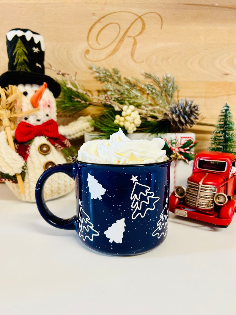 May include: A blue ceramic mug with white Christmas trees and white speckles. The mug is filled with whipped cream.