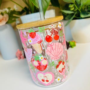 Peut inclure: Gobelet en verre transparent avec un couvercle en bambou et une paille en verre transparent. Le gobelet est d&eacute;cor&eacute; d'illustrations roses et rouges de fraises, de cerises, de cr&egrave;me glac&eacute;e et de coquillages. Le design comprend des marguerites blanches et un c&oelig;ur avec des cerises.