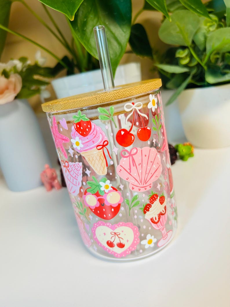 Peut inclure: Gobelet en verre transparent avec un couvercle en bambou et une paille en verre transparent. Le gobelet est d&eacute;cor&eacute; d'illustrations roses et rouges de fraises, de cerises, de cr&egrave;me glac&eacute;e et de coquillages. Le design comprend des marguerites blanches et un c&oelig;ur avec des cerises.