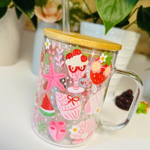 Peut inclure: Une tasse en verre transparent avec un couvercle en bambou et une paille transparente. La tasse est d&eacute;cor&eacute;e d'illustrations roses et rouges sur le th&egrave;me de l'&eacute;t&eacute;, notamment des fraises, des coquillages et une &eacute;toile de mer. La tasse a une poign&eacute;e transparente.
