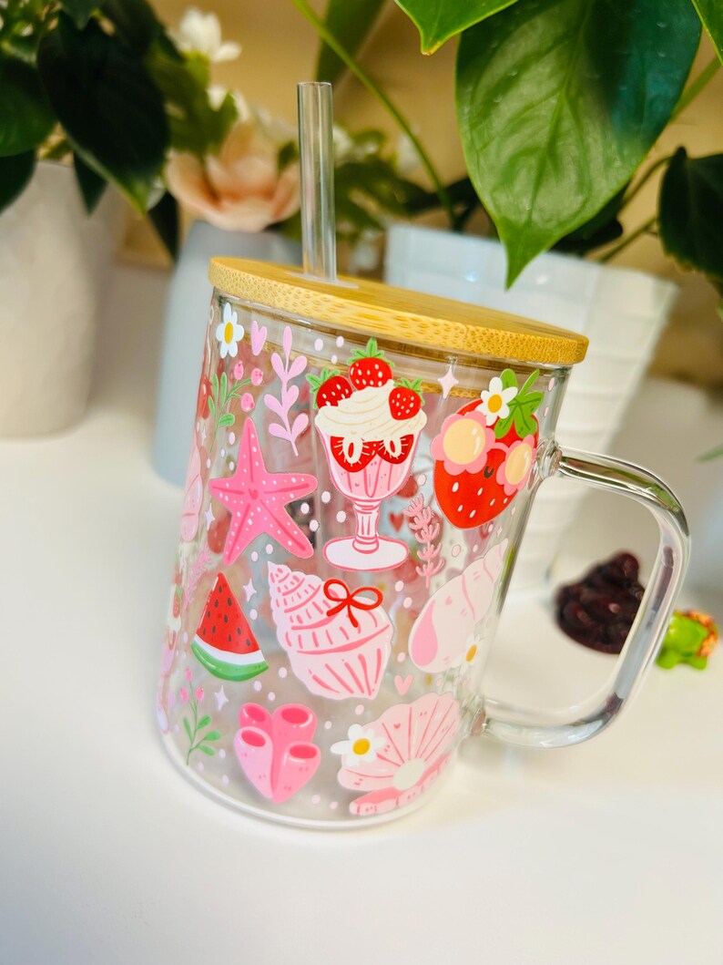 Peut inclure: Une tasse en verre transparent avec un couvercle en bambou et une paille transparente. La tasse est d&eacute;cor&eacute;e d'illustrations roses et rouges sur le th&egrave;me de l'&eacute;t&eacute;, notamment des fraises, des coquillages et une &eacute;toile de mer. La tasse a une poign&eacute;e transparente.