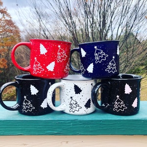 May include: Four ceramic mugs with a speckled glaze and a white outline of a Christmas tree design. The mugs are stacked in a pyramid shape and are in the colors red, navy blue, white, and black.