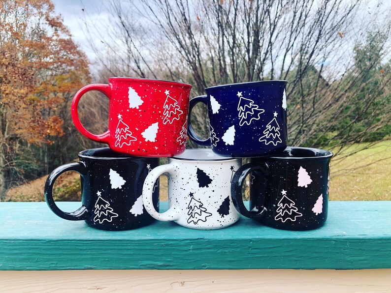 May include: Four ceramic mugs with a speckled glaze and a white outline of a Christmas tree design. The mugs are stacked in a pyramid shape and are in the colors red, navy blue, white, and black.