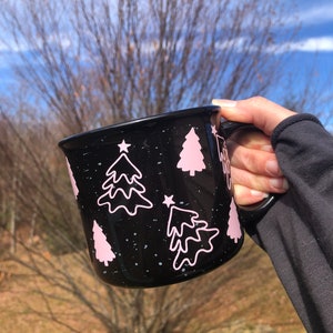 May include: Black ceramic mug with a white speckled glaze and pink Christmas tree designs. The mug has a white star on each tree.