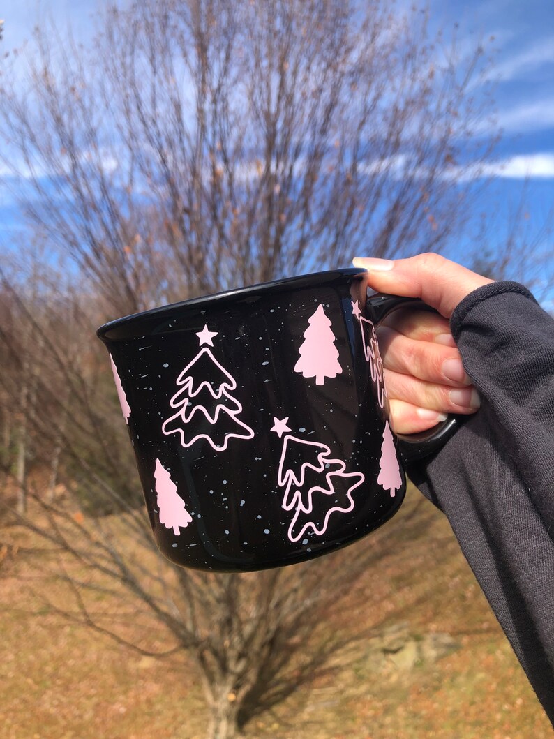 May include: Black ceramic mug with a white speckled glaze and pink Christmas tree designs. The mug has a white star on each tree.