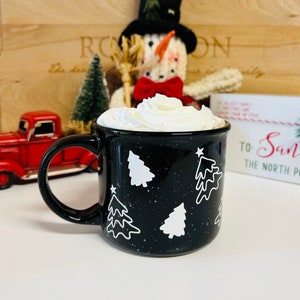 May include: Black ceramic mug with white Christmas tree designs and white speckles. The mug is filled with whipped cream and is sitting on a white surface. The mug says "To Santa The North Pole" on the side.