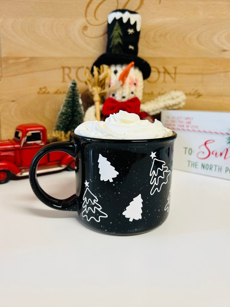May include: Black ceramic mug with white Christmas tree designs and white speckles. The mug is filled with whipped cream and is sitting on a white surface. The mug says "To Santa The North Pole" on the side.