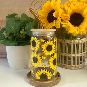 May include: A glass jar with a wooden lid and a gold metal straw. The jar is decorated with a sunflower pattern and has a wooden coaster underneath.