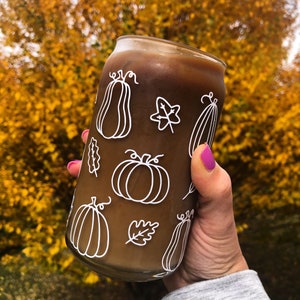 May include: A brown iced coffee drink in a reusable glass can with a white design of pumpkins and leaves. The can is being held by a hand with pink nail polish.