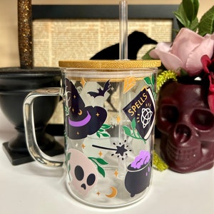 May include: A clear glass mug with a bamboo lid and a straw. The mug features a Halloween theme with illustrations of a witch's hat, a cauldron, a skull, a wand, and a book that says "Spells".