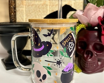 May include: A clear glass mug with a bamboo lid and a straw. The mug has a Halloween theme with illustrations of a witch's hat, a cauldron, a skull, a wand, and a book that says "Spells".
