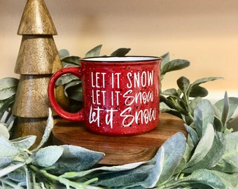May include: Red ceramic mug with white speckled glaze and the text "Let it snow" repeated three times. The mug is sitting on a wooden coaster surrounded by a faux greenery garland.