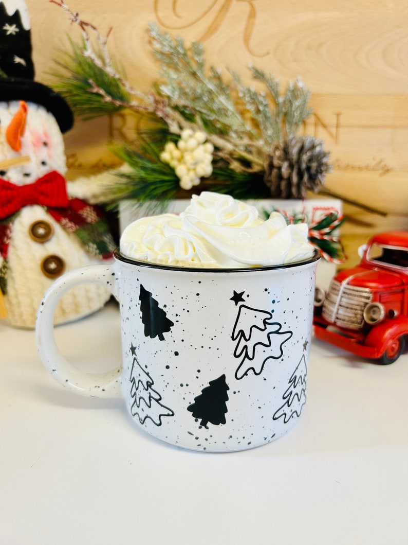 May include: White ceramic mug with black and white Christmas tree designs and black speckles. The mug is filled with a white whipped cream topping.