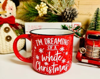 May include: Red ceramic mug with a white speckled glaze. The mug has a black rim and handle. The mug says "I'm Dreaming of a White Christmas" with snowflakes around the text.