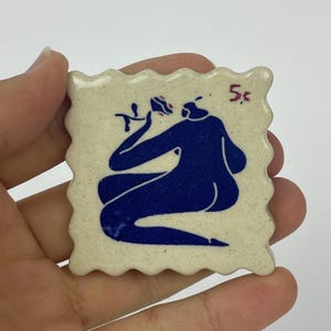 May include: A square, off-white ceramic piece with a wavy edge, featuring a blue illustration of a figure and a flower. The number "5¢" is written in red. The piece is held in a hand.
