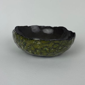 May include: A small, handmade ceramic bowl with a textured, green exterior and a dark interior. The bowl's rim has an irregular, organic shape. The green surface has a speckled pattern, adding visual interest to the piece. The bowl is likely used for serving or display.