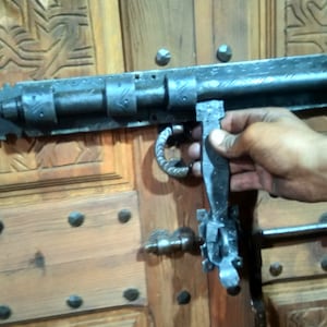 May include: A black metal door latch with a decorative design. The latch is attached to a wooden door with a carved pattern.