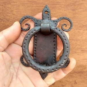 Peut inclure: Un heurtoir de porte en métal noir avec une poignée en forme d'anneau et des volutes décoratives. Le heurtoir a une finition martelée et une surface texturée.