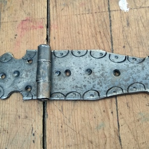 May include: A weathered, metal hinge with a decorative design. The hinge is gray with a distressed finish, featuring circular and curved cutouts along its length. It has multiple holes for mounting and a central pivot.