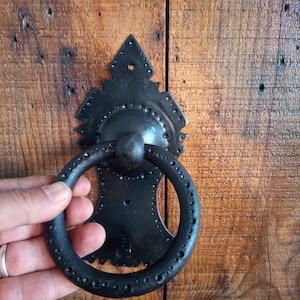 Könnte beinhalten: Ein schwarzer Türklopfer aus Metall mit einem Ringgriff und dekorativen Details. Der Türklopfer ist an einer Holztür montiert.