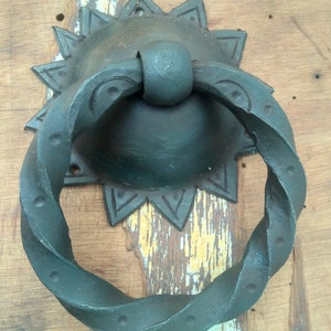 Può includere: Un batacchio per porte in metallo nero con un design a corda attorcigliata e una base a forma di stella. Il batacchio è montato su una porta in legno.